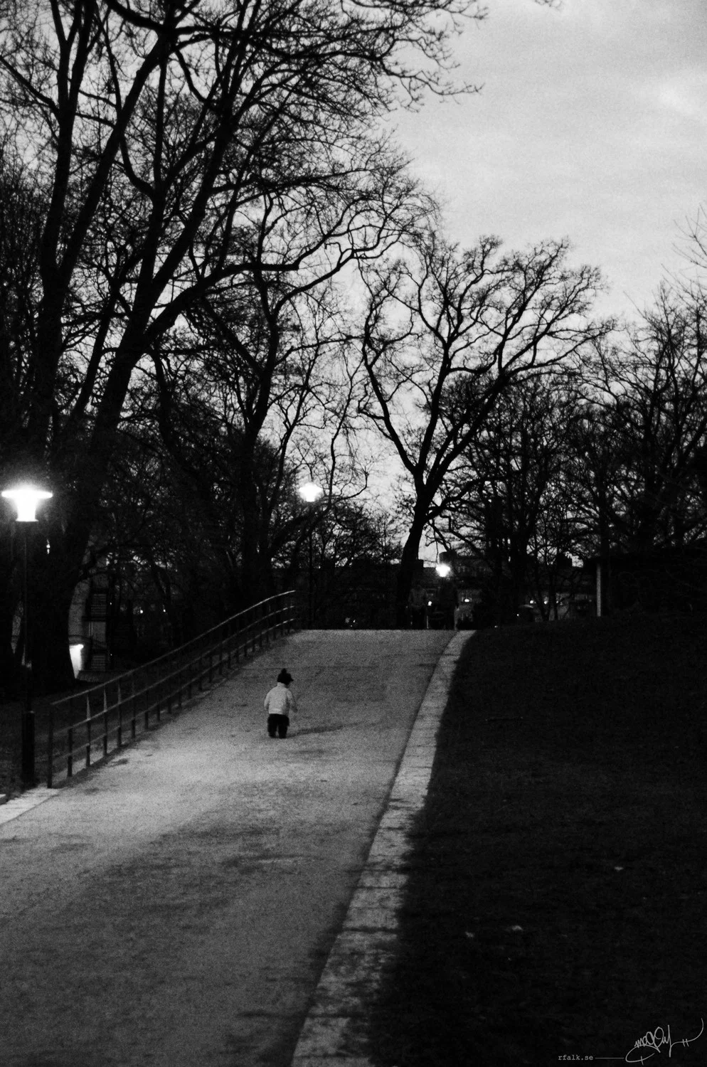 A black-and-white photograph of a wide, gently rising park path at dusk, lined with tall, leafless trees. A small figure in a light-coloured jacket walks away from the camera near the centre of the path. Illuminated street lamps stand among the trees, while a metal railing follows the left side and a dark grassy slope borders the right.