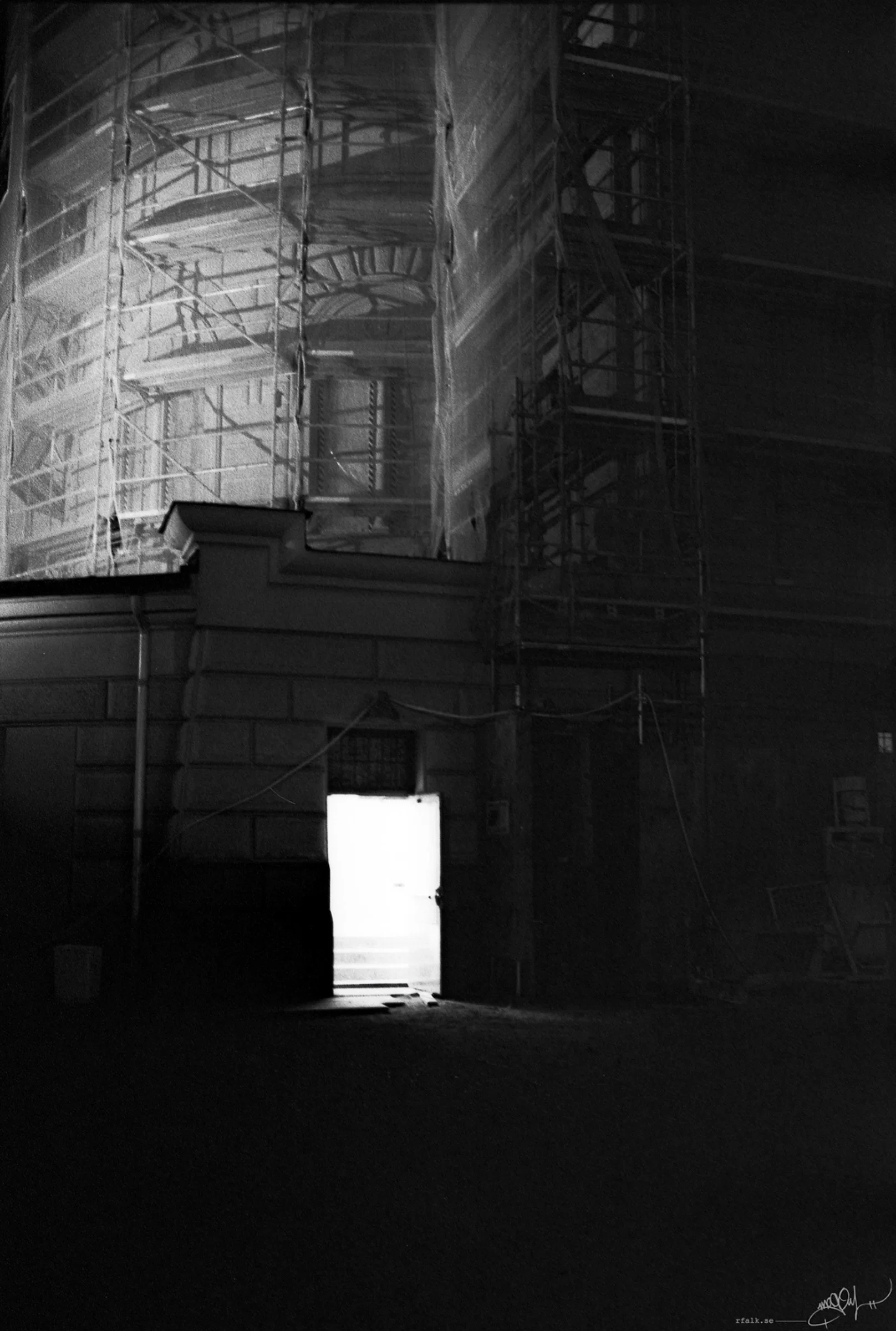 A black-and-white nighttime photograph of a large building covered in scaffolding and translucent protective sheeting. The upper left section is brightly illuminated, revealing a dense structure of metal poles and platforms, while the right side disappears into darkness. Near the bottom centre, an open doorway emits a bright rectangular area of light onto the otherwise dark ground.