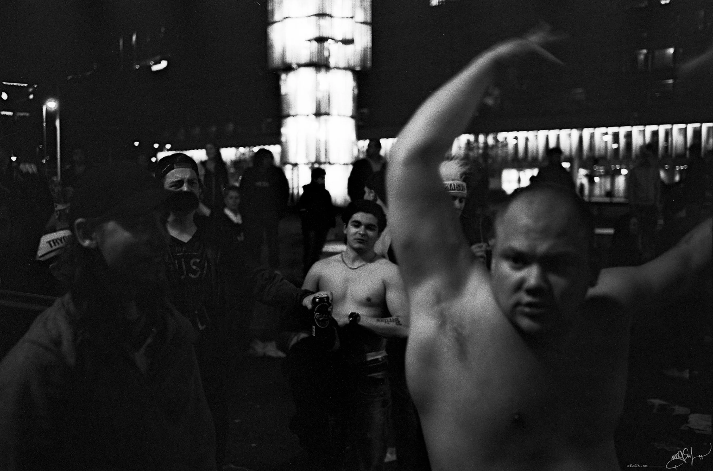 A black-and-white nighttime street photograph of a group of people gathered in a public square. A shirtless man dominates the right foreground with both arms raised above his head, while another shirtless person stands near the centre holding a can. Several other figures occupy the darker background, with an illuminated building façade and a tall glowing column rising behind them.