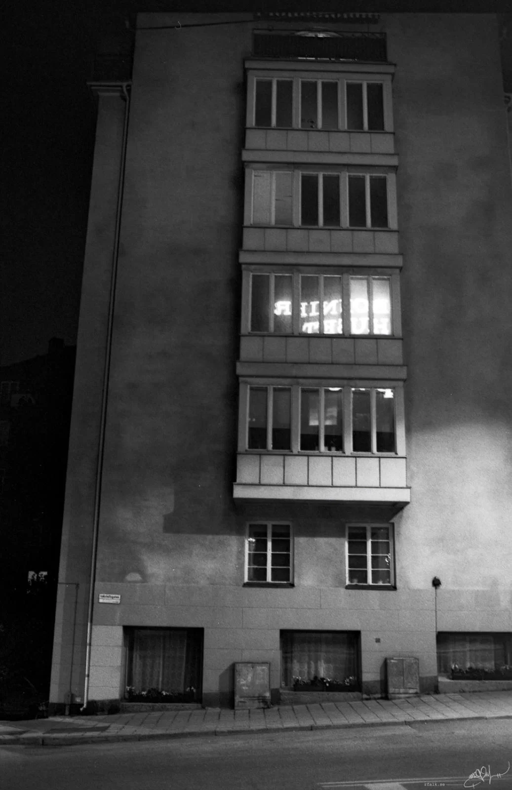 A black-and-white nighttime photograph of a tall residential building façade viewed from across an empty street. A vertical stack of enclosed balconies runs up the centre of the building, with most windows dark. Bright, partially obscured lettering shines through one of the middle windows, while a strong light illuminates part of the wall on the right. The pavement and ground-floor windows are visible below.