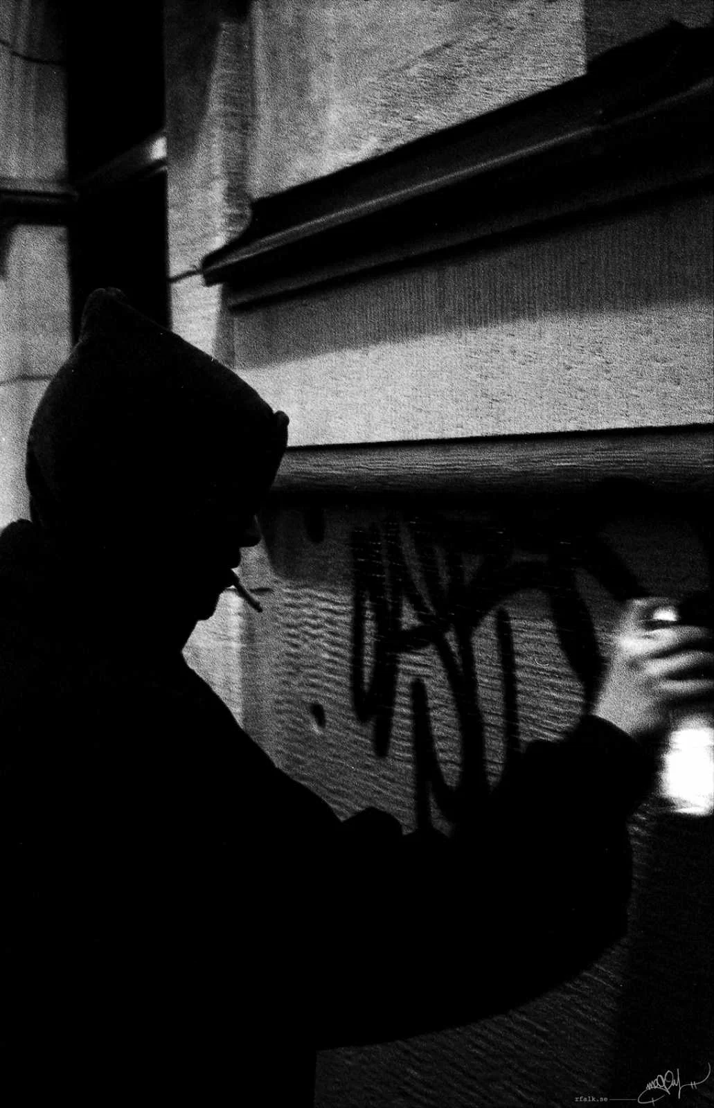A grainy black-and-white photograph of a hooded person in profile, standing close to a wall covered with graffiti. A cigarette protrudes from the person's mouth, while one raised hand holds a spray-paint can near the right edge of the frame. The figure appears almost entirely as a dark silhouette against the lighter, textured building façade.
