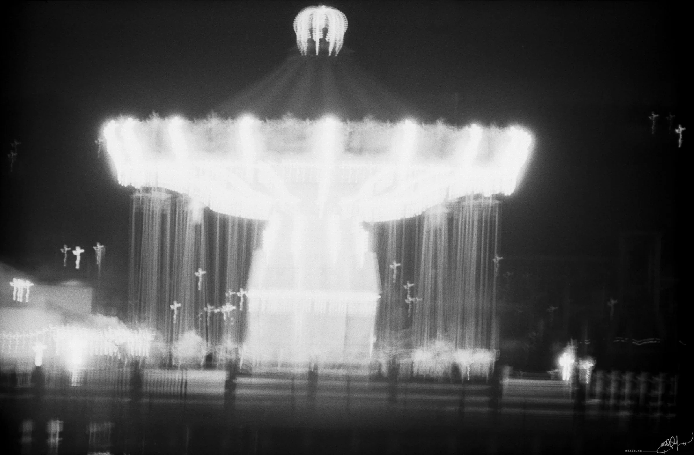 A black-and-white nighttime photograph of a brightly illuminated swing carousel at an amusement park. The circular canopy and long chains of the ride are blurred by motion and overexposure, creating vertical streaks of white light against the dark sky. Smaller illuminated structures and faint silhouettes are visible near the ground.