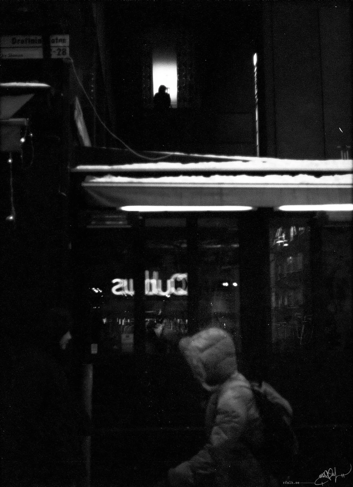 A black-and-white nighttime street photograph of a hooded person walking across the lower foreground, carrying a dark backpack. Behind the figure, a brightly lit horizontal awning separates a glass storefront from the darker building above. Reversed illuminated lettering is visible through the glass, while a small silhouetted figure appears in a brightly lit upper window.