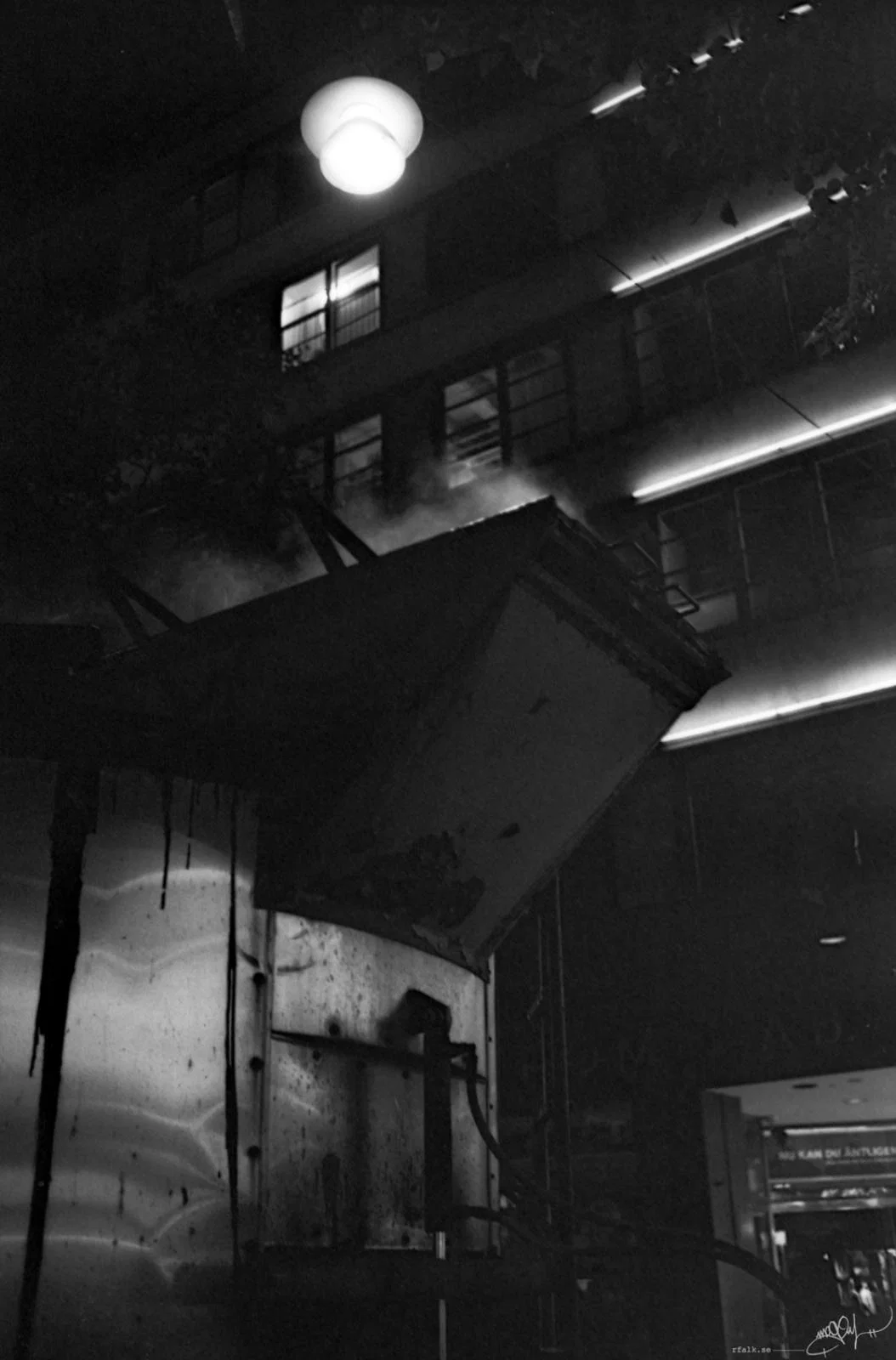 A black-and-white nighttime photograph looking upward at a building façade with illuminated windows and horizontal strips of light. A large angular metal structure projects diagonally across the foreground, with steam or mist rising behind it. A bright spherical street lamp hangs near the top of the frame, while part of a lit entrance is visible in the lower right corner.