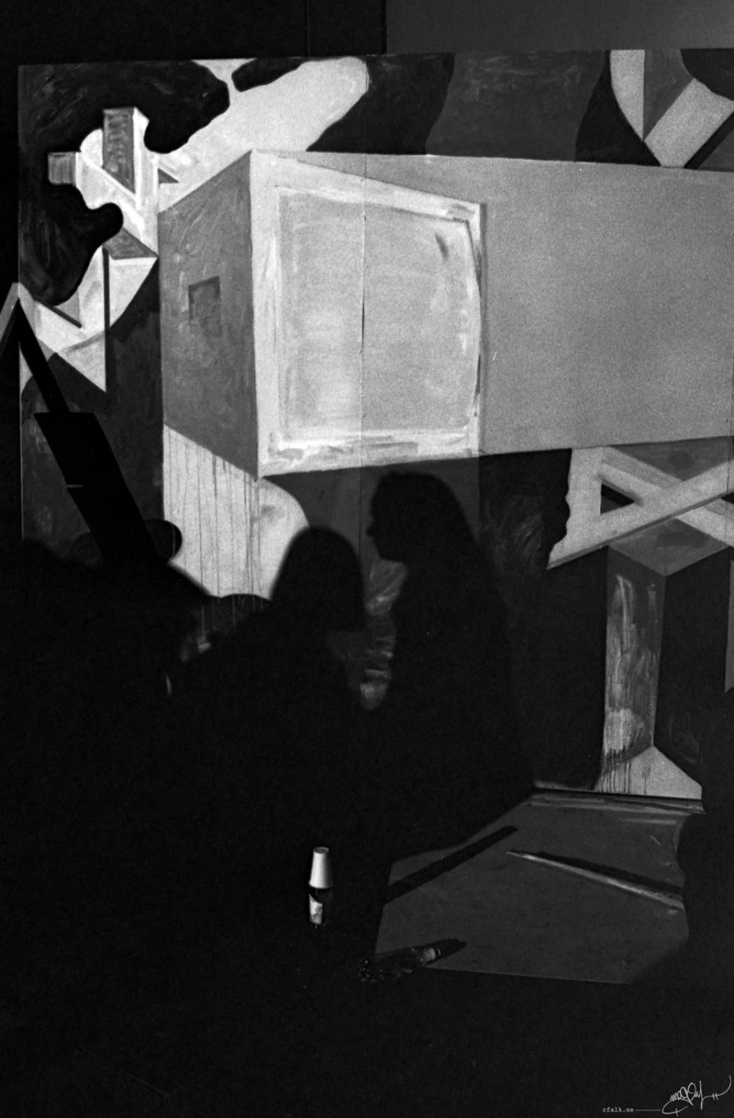 A black-and-white photograph of several people standing in front of a large geometric mural. Their dark silhouettes overlap across the lower half of the image, while the mural behind them features angular shapes, intersecting planes and a prominent rectangular form resembling an open box. A table or counter occupies the lower right foreground.
