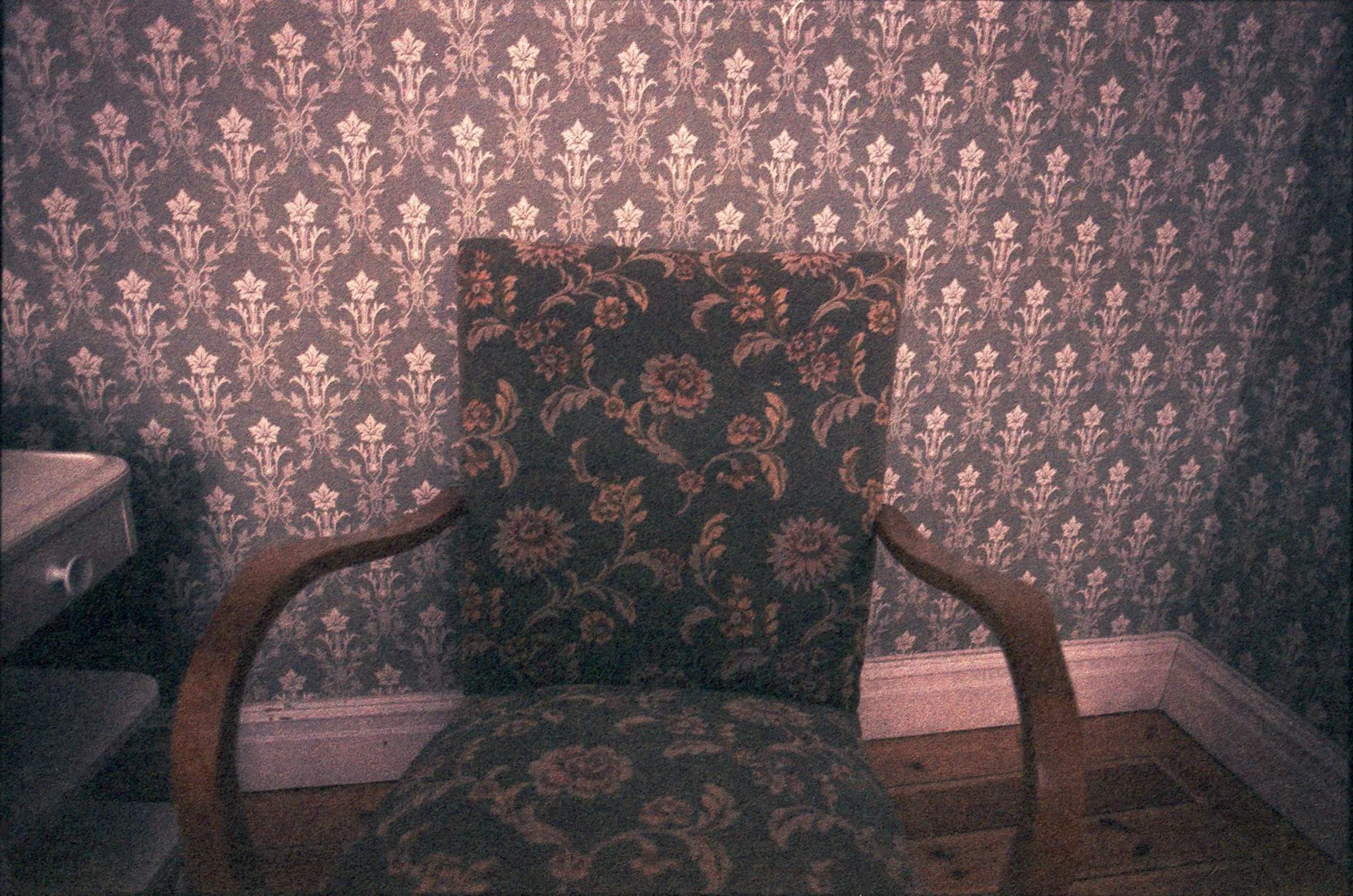 An upholstered armchair with curved wooden armrests stands against floral-patterned wallpaper. The chair's darker floral fabric contrasts with the smaller repeating pattern on the wall, in muted pink and brown tones.