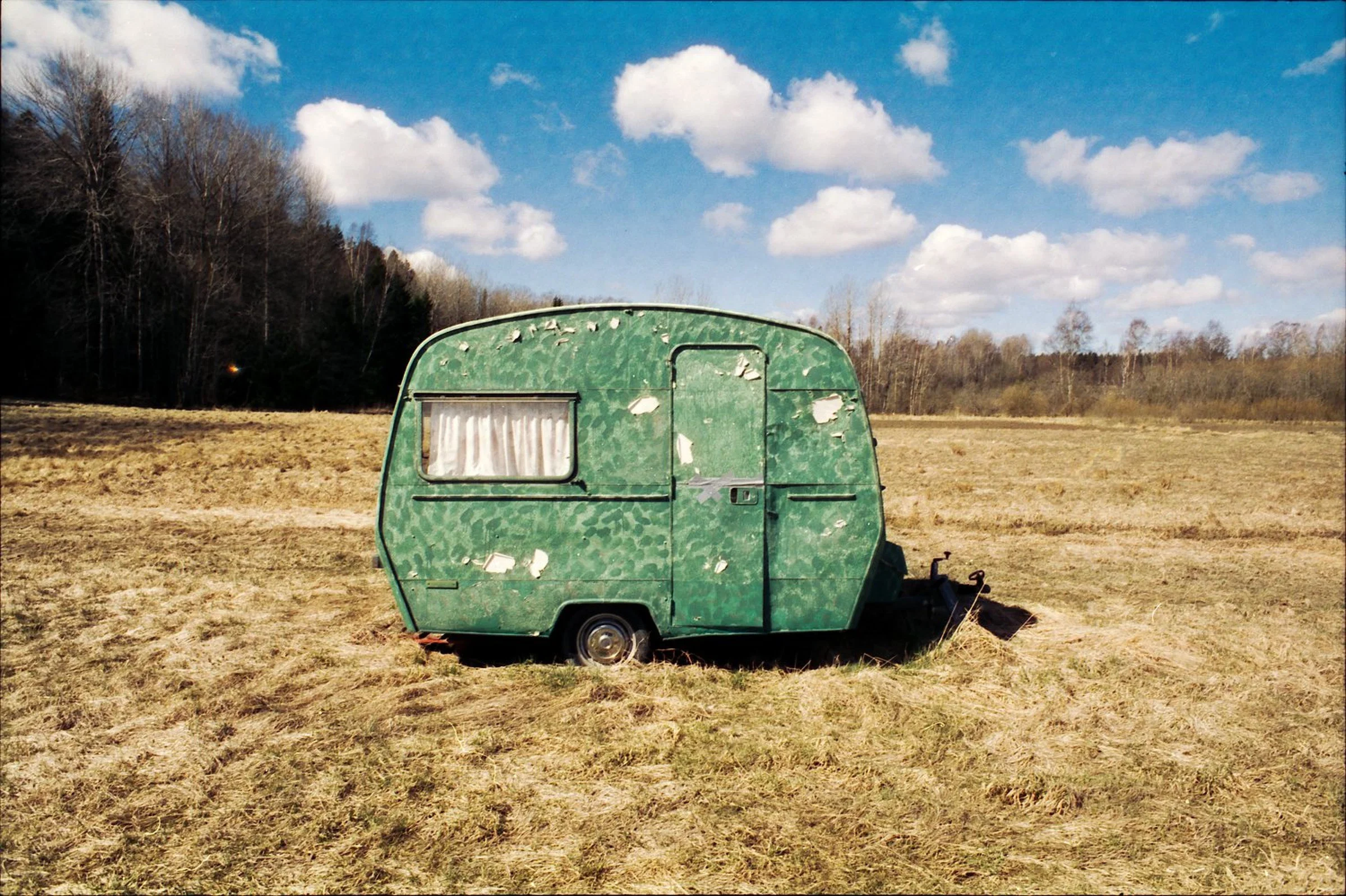 A small green caravan with peeling paint stands alone in a field of dry, yellow-brown grass. A line of trees borders the field beneath a bright blue sky with scattered white clouds.