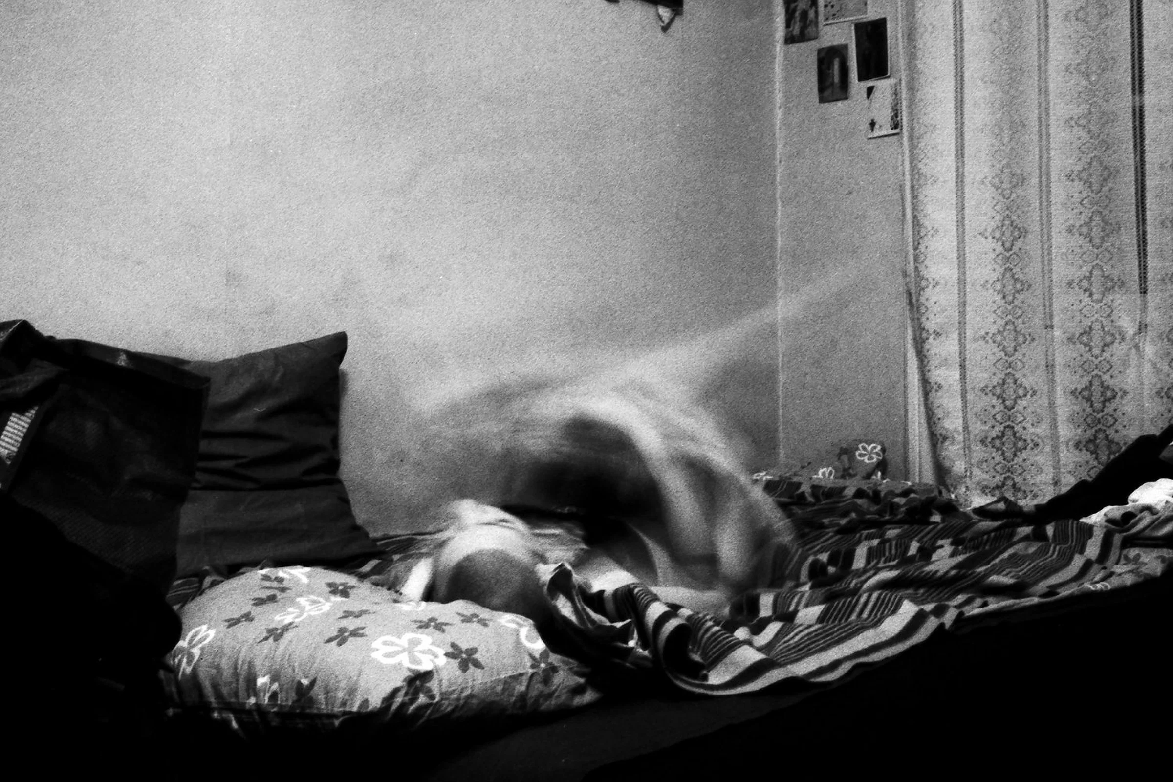 A black-and-white photograph of a person lying on an unmade bed, their head visible while their upper body is blurred by movement. Patterned bedding, dark pillows and lace curtains surround the figure in a small bedroom.
