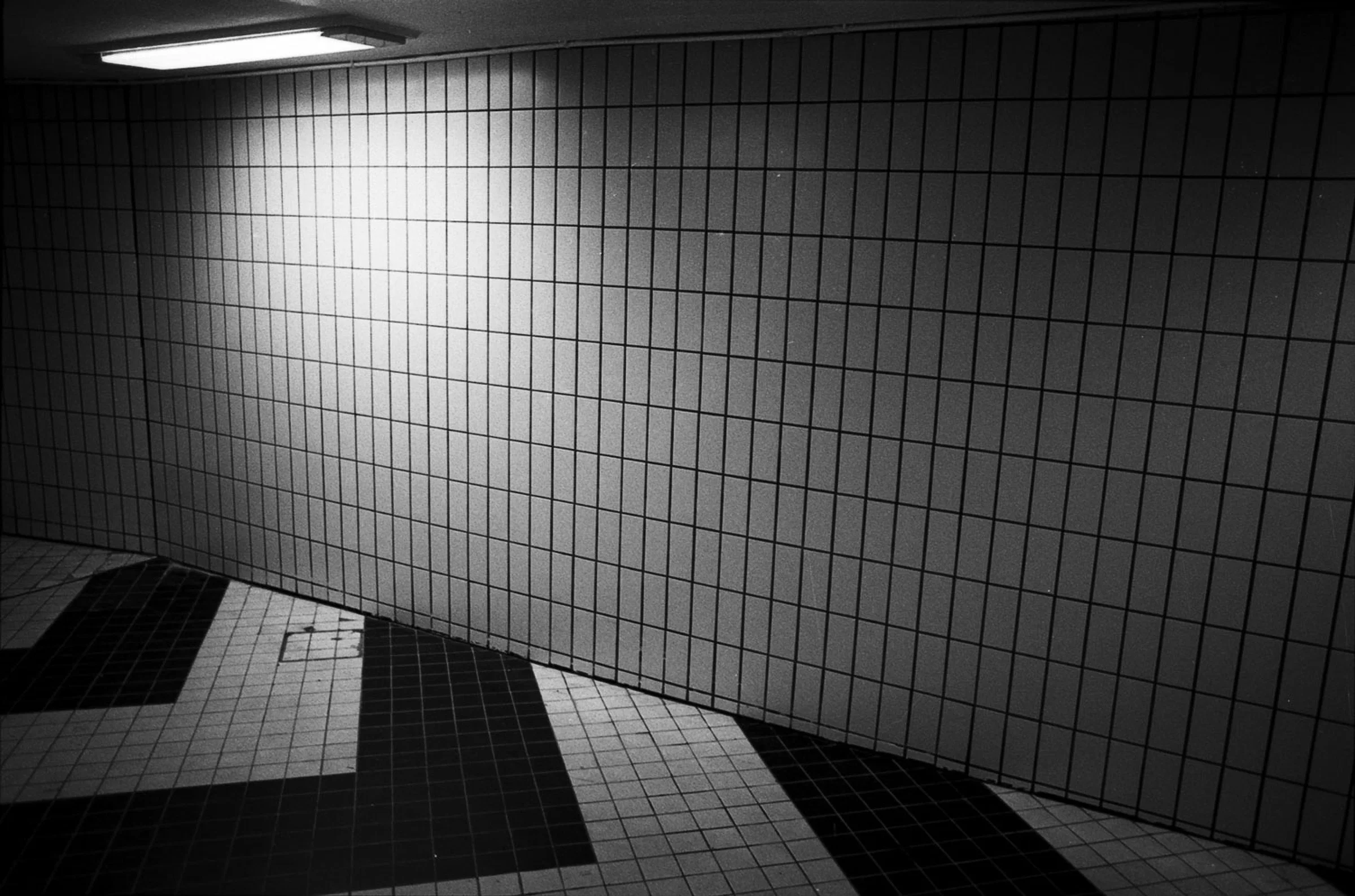 A black-and-white photograph of an empty tiled interior, with a long wall of square tiles extending diagonally across the frame. A rectangular fluorescent ceiling light illuminates part of the wall, while large geometric areas of dark and light tiles form a pattern across the floor in the foreground.
