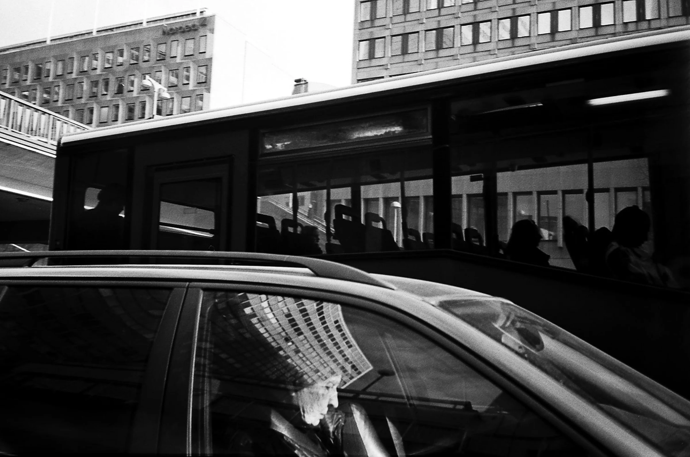 A black-and-white street photograph showing a bus passing behind a car in the foreground. Dark silhouettes of passengers are visible through the bus windows, while the car's side window reflects the grid of a nearby building. Office buildings rise in the background, creating overlapping layers of windows, reflections and figures.