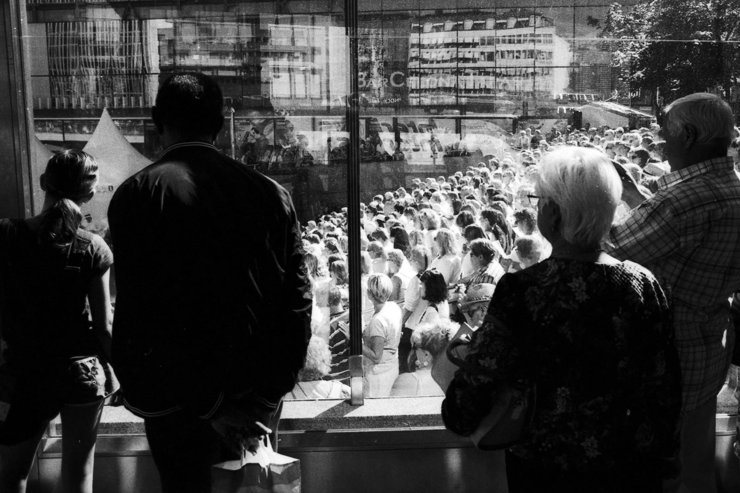 A black-and-white photograph of four people standing with their backs or sides to the camera, looking through a large glass window at a densely packed crowd outside. Reflections of surrounding buildings and the crowd overlap across the glass, while the people in the foreground appear as dark silhouettes against the brightly lit gathering.