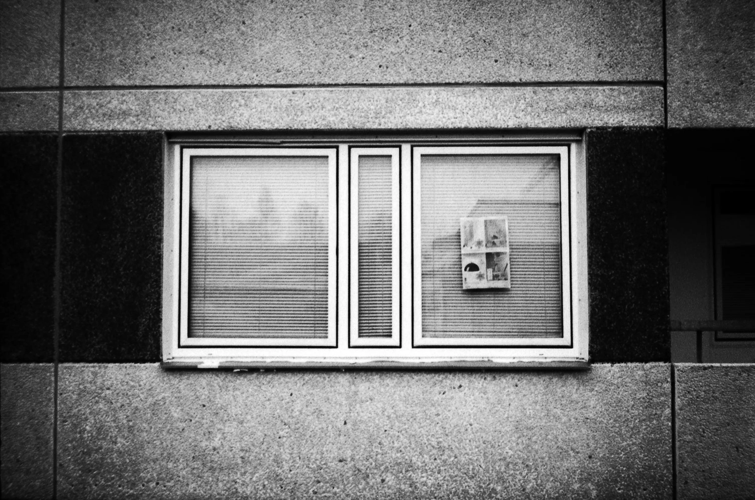 A black-and-white photograph of a rectangular window set into a concrete building façade. Three sections of the window are framed in white, with closed horizontal blinds behind the glass. A small paper notice with several images is attached to the right-hand pane. Dark horizontal bands in the surrounding wall contrast with the lighter concrete panels.