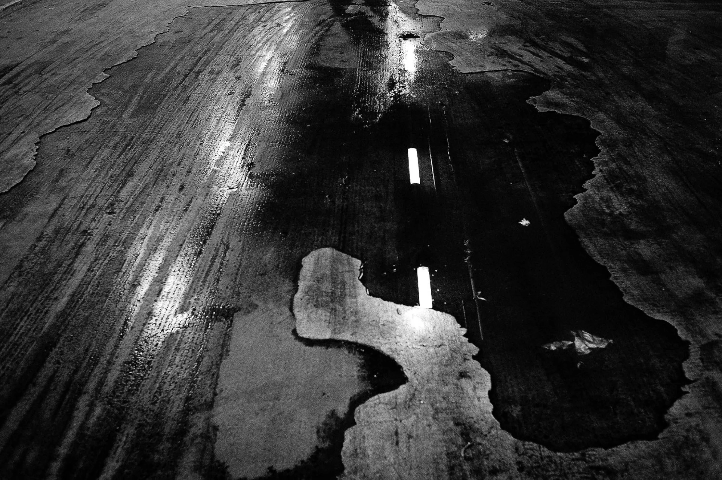 A black-and-white photograph looking down at a wet, worn road surface. An irregular dark puddle extends through the centre and right side of the frame, reflecting two bright rectangular lights. Long streaks, cracks and patches of moisture run diagonally across the surrounding surface, creating contrasting areas of light and shadow.