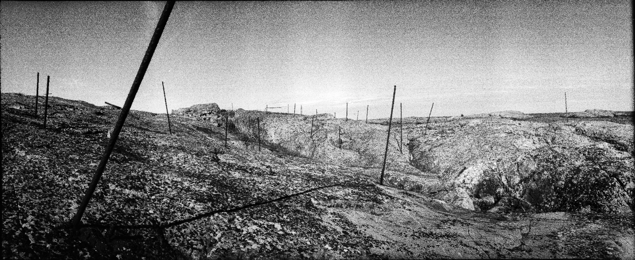 A grainy black-and-white panorama of bare, rocky ground crossed by thin, leaning fence posts. A large dark post cuts diagonally across the left foreground beneath an open sky.
