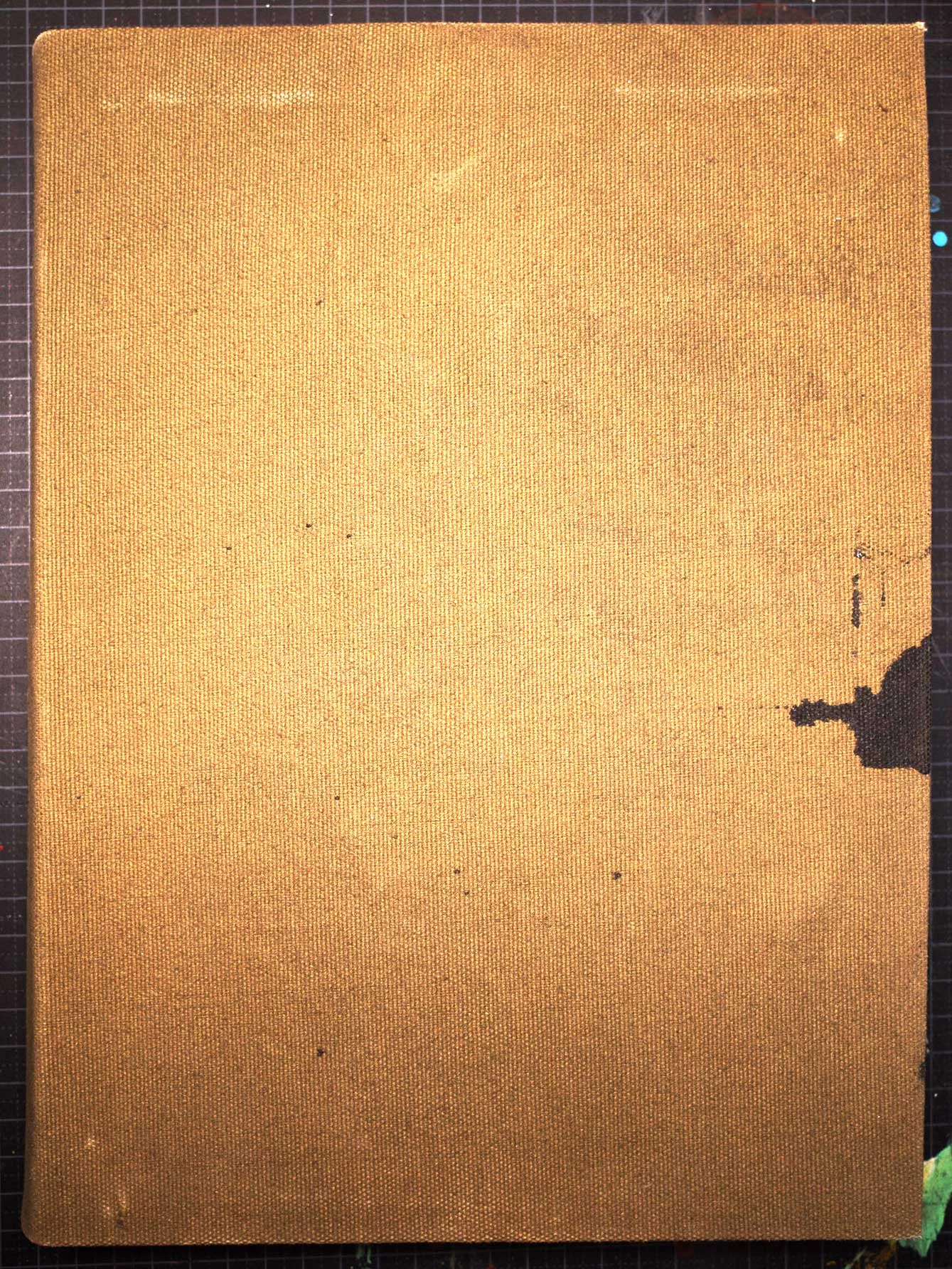 A photograph of a closed sketchbook with a plain ochre-brown fabric cover, lying on a dark gridded cutting mat. The cover has rounded corners, small scattered marks and a dark worn patch along its right edge.