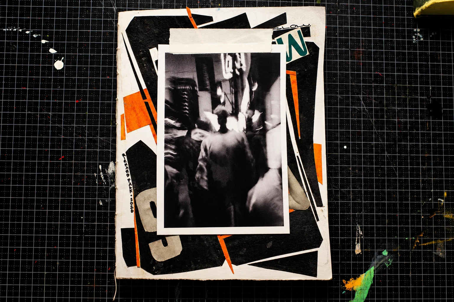 A handmade book cover on a black gridded cutting mat, with black, orange and white collage shapes beneath a taped black-and-white street photograph of people moving through a crowd.