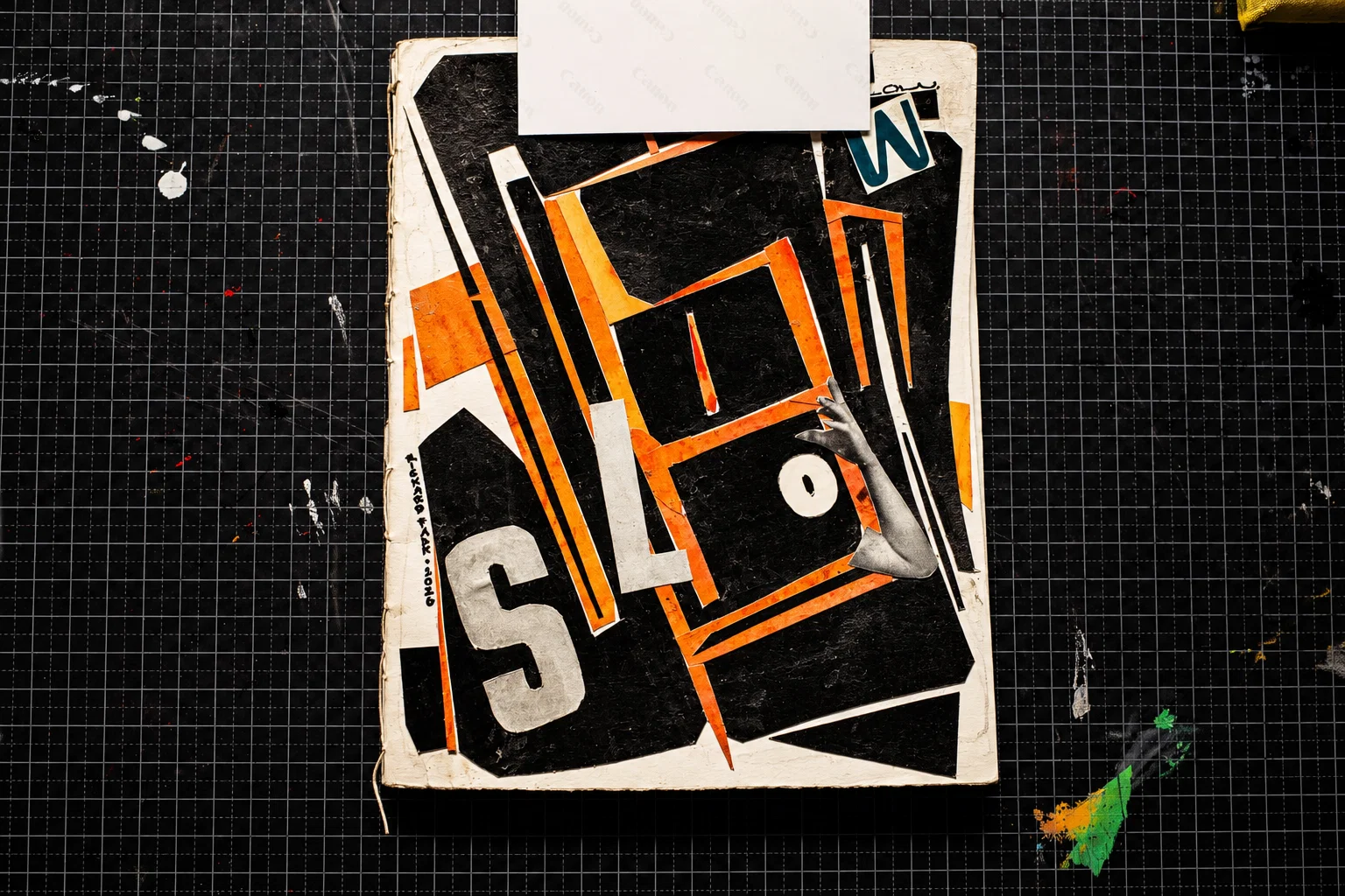 A handmade book cover with large, overlapping letters spelling SLOW, arranged among angular black, orange and white shapes. A cut-out photographic arm extends across the lettering. The book rests on a black gridded cutting mat.
