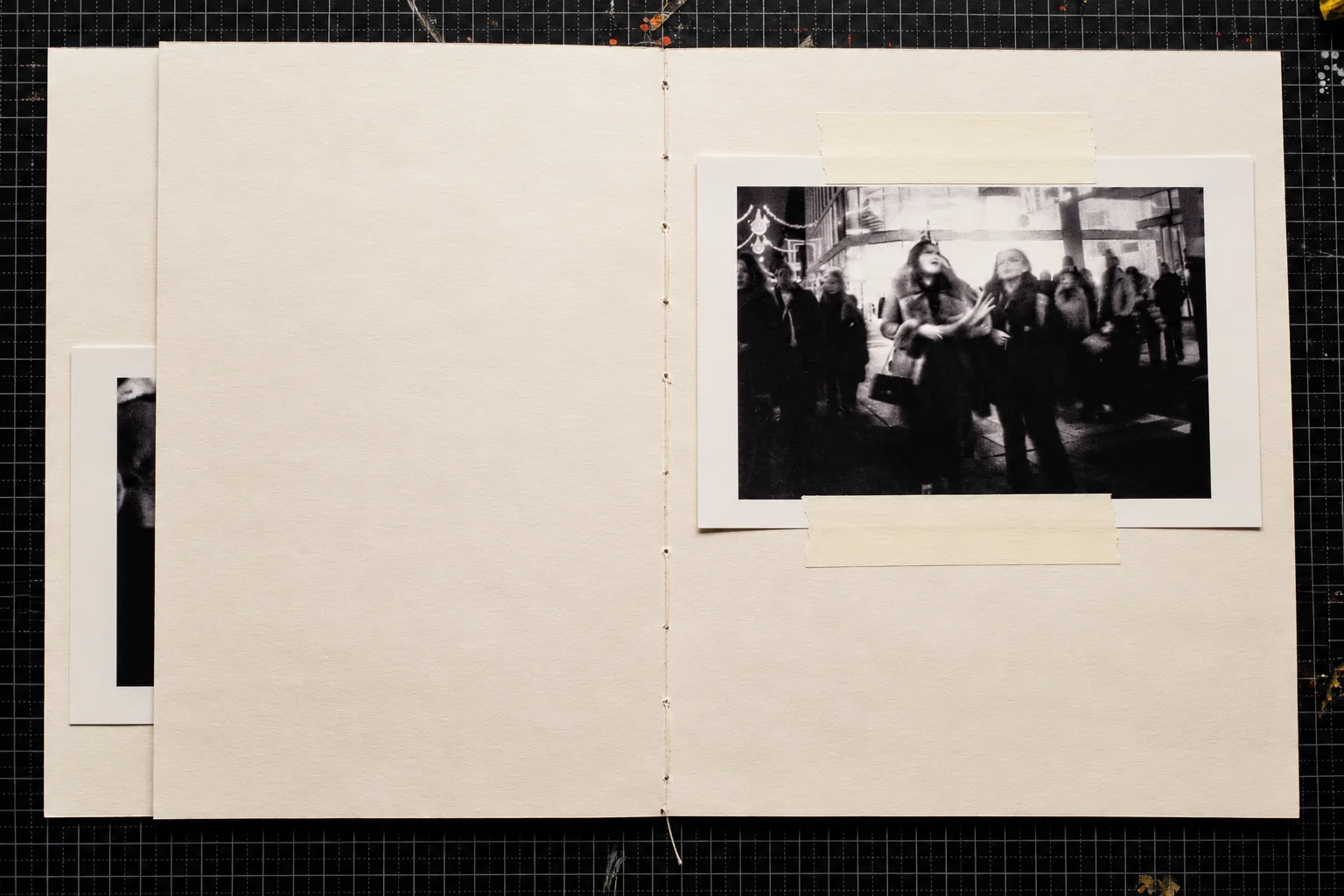 A single black-and-white photograph on the right-hand page shows people standing and walking along a busy pedestrian street. Two figures near the centre are more clearly visible against the surrounding crowd. The left page is blank.