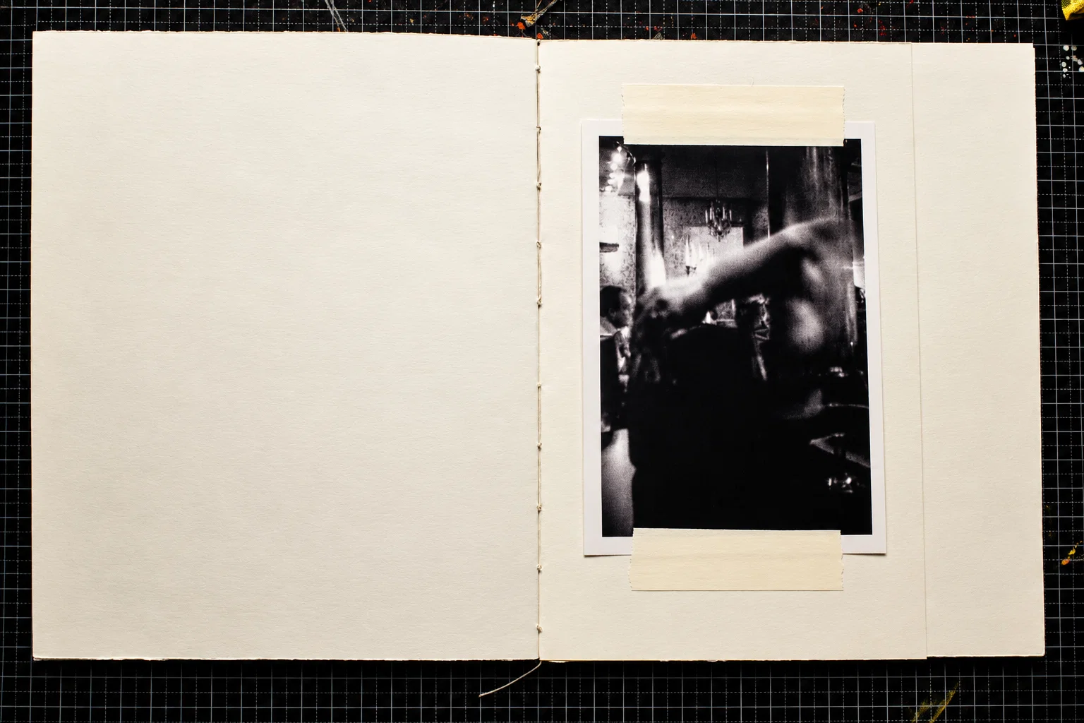 A single black-and-white photograph on the right-hand page shows a dimly lit interior with people gathered beneath hanging lights. A large, blurred figure and an outstretched arm dominate the foreground. The left page is blank.